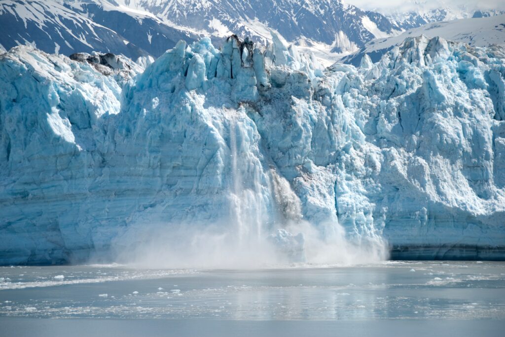Blue glacier with ice falling into the water below and mountains in the background.