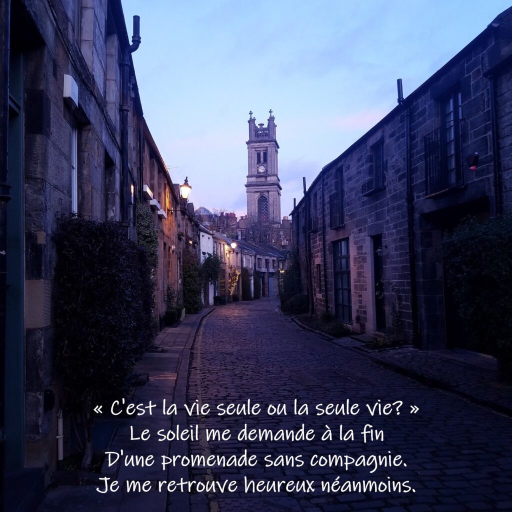 Dimly lit street at dusk with brick buildings on each side and a clock tower at the end of the street. Text on photo reads: "C'est la vie seule ou la seule vie? Le soleil me demande à la fin. D'une promenade sans compagnie. Je me retrouve heureux néanmoins."