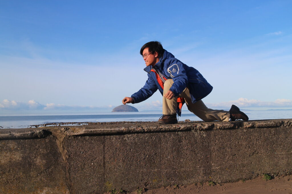 Derek bent over as if throwing a curling stone. In the background you can see the island of Ailsa Craig which is shaped in a half dome like a curling stone.