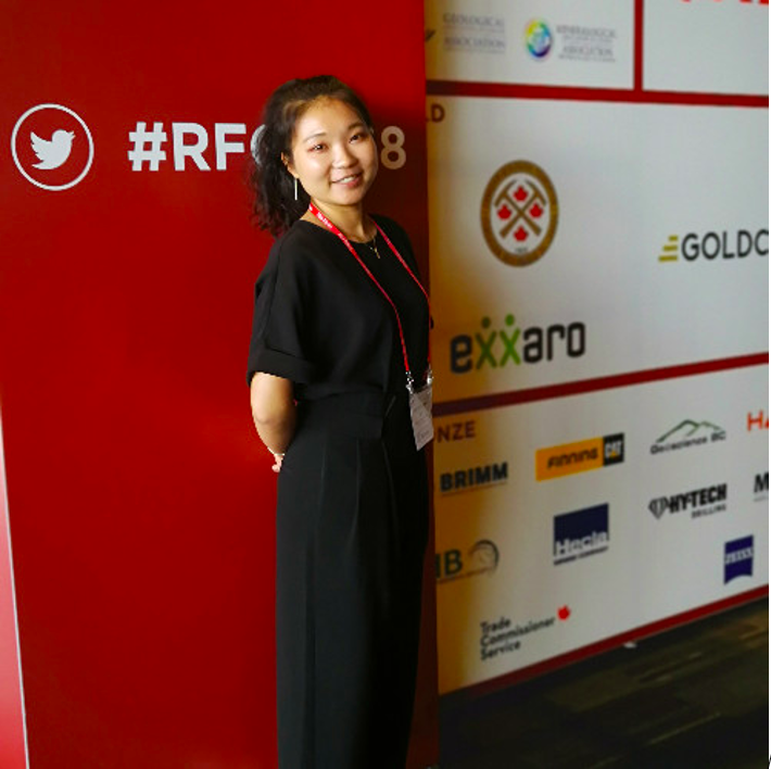 Woman in a long black dress standing with her hands behind her back and smiling at the camera. She is standing in front of a red sign with symbols on it.