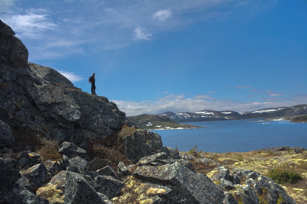 person in the distance stands on a large mound of rocks overlooking a body of water with mountains in the background.