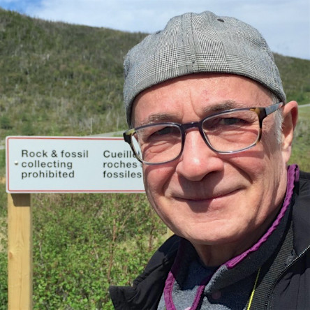 Man smiles at the camera wearing large square glasses and a grey hat. Behind him there is a sloping green hill and a sign that reads: "Rock and fossil collecting prohibited."