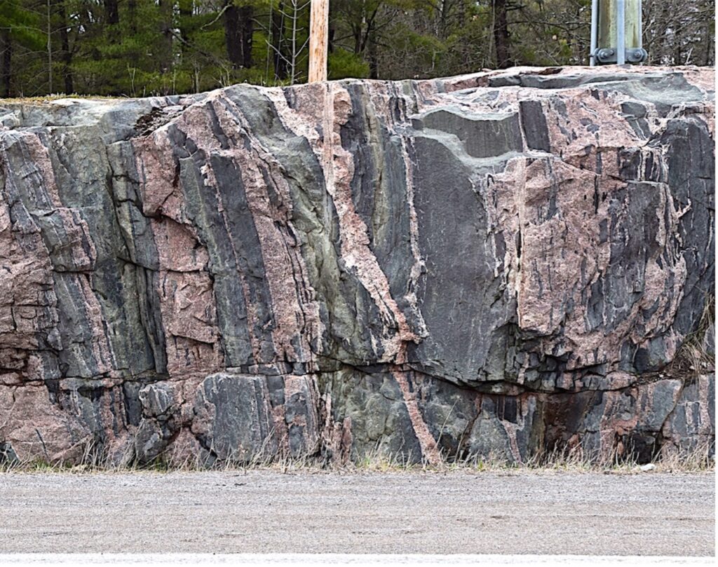 Rock outcrop on the side of a road. Black and pink rock alternate in roughly vertical lines