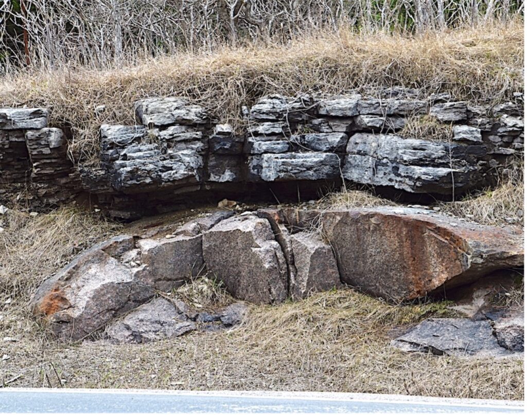 Rock outcrop on the side of a road. The top rock layer is a darker colour with defined layers. The bottom rock is lighter pink and curves downwards at the sides of the photo