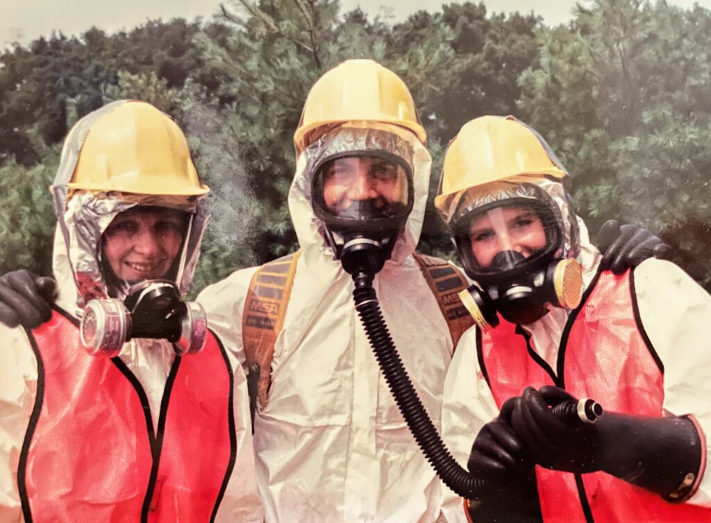 Three people smiling at the camera. They are dressed in full PPE with helmets, gas masks and full suits.