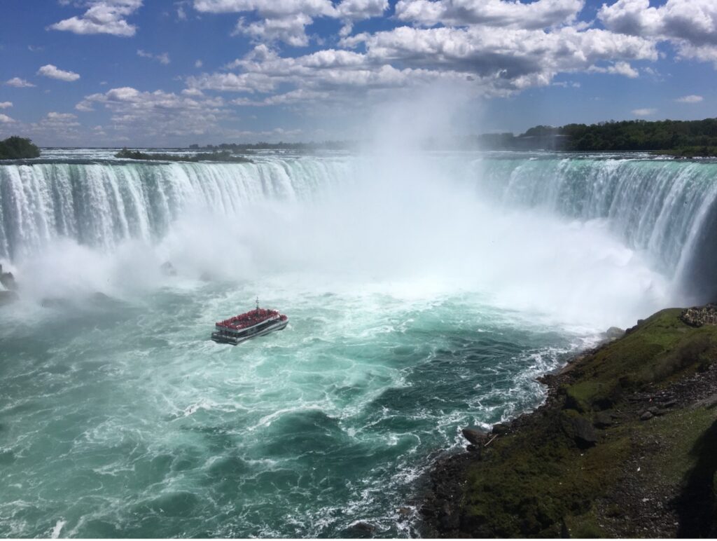 View of Niagara Falls from the Northern (Canadian) side. The sun is shining and the water is teal blue. A boat can be seen going toward the falls.