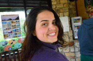 Girl looking over her shoulder smiling at the camera. She has brown hair and is wearing a purple sweater.
