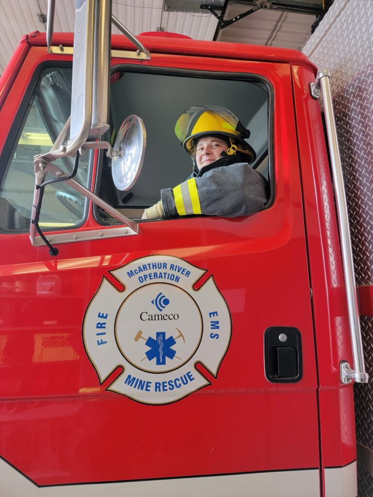 A woman sitting in the drivers seat of a fire truck wearing a firefighting suit and smiling at the camera. The left-side door of the truck can be seen with the Cameco logo on it.