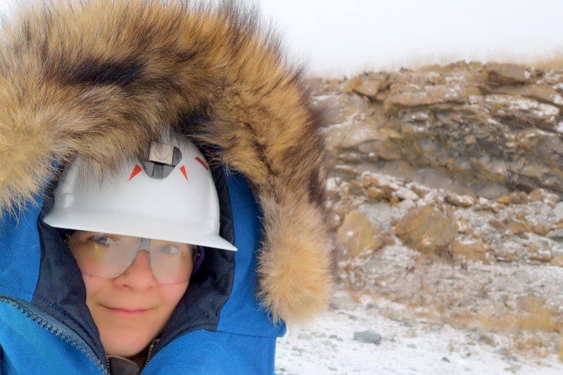 A woman smiling at the camera wearing a white helmet, safety glasses, and a thick blue winter jacket with a fur lined hood. In the background there are brown rocks with a bit of snow on them.