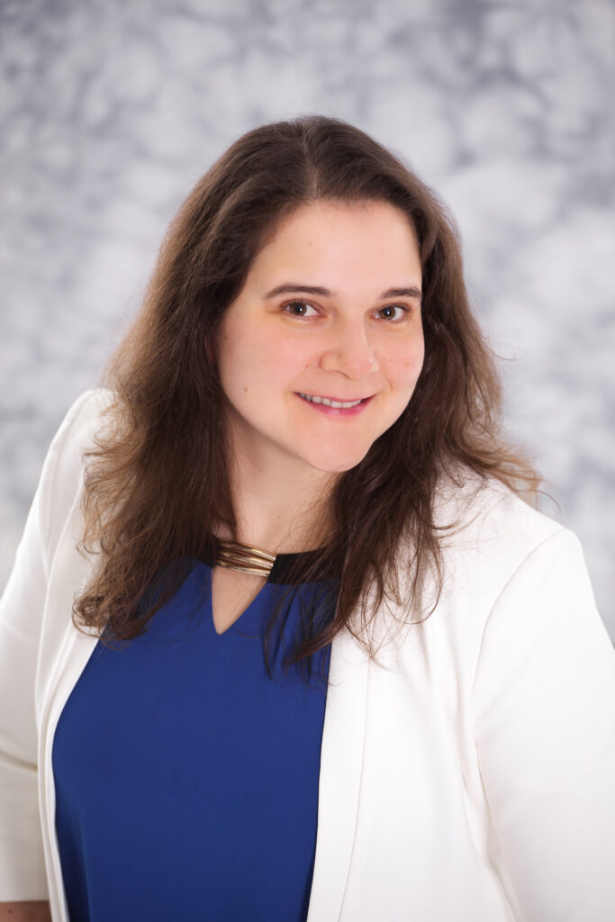 Woman with brown hair wearing a blue shirt and white blazer smiles at the camera. Behind her there is a grey blurred photo background.