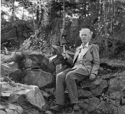 Black and white photo of an elderly woman sitting on a rock holding a rock hammer in one hand and smiling into the distance.