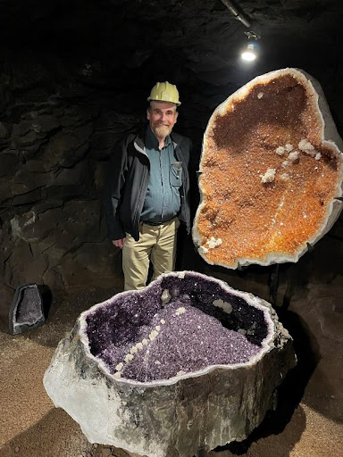 A man in a hard hat stands next to a large clamshell shaped amethyst geode.