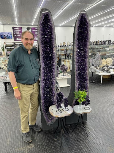 A an in a green shirt stands next to two tall rectangular-shaped amethyst geodes. The geodes are taller than the man.