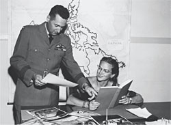 A black and white photograph of a woman sitting at a desk holding an open book. A man stands beside her and points to something in the book while holding a piece of paper in his other hand. There is a map of Canada on the wall behind them.