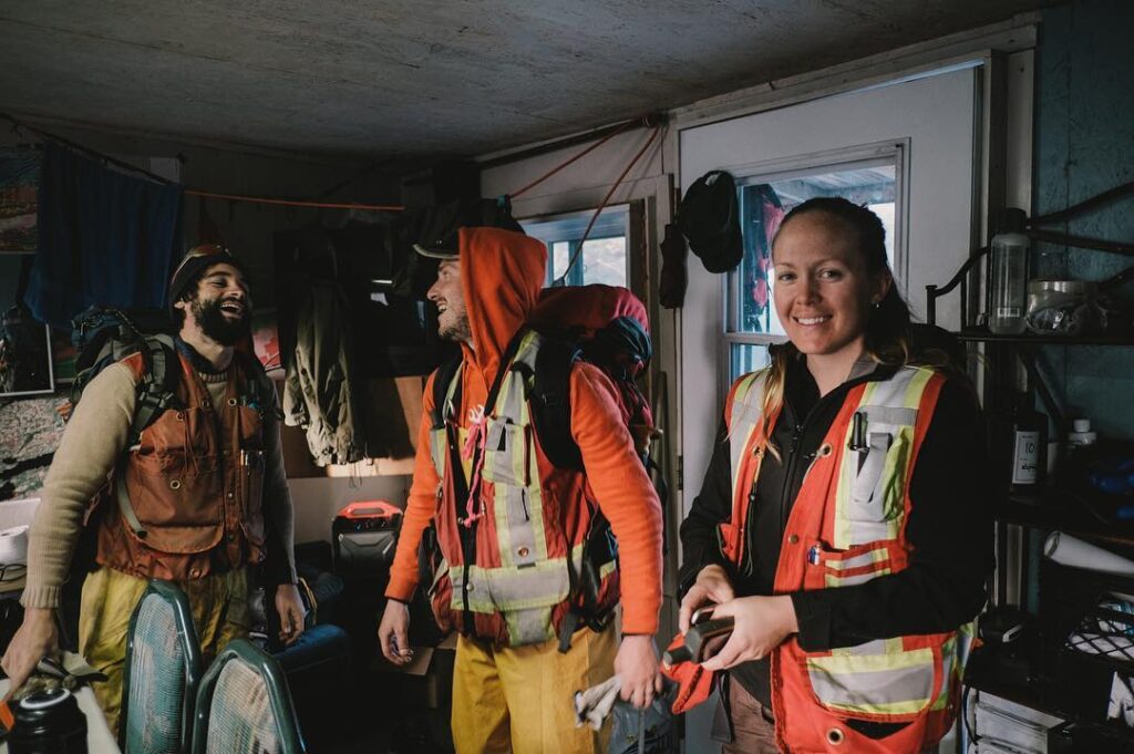 Three people wearing red safety jackets. The closest to the camera is smiling at the camera, while the other two people are looking at one another and appear to be laughing.