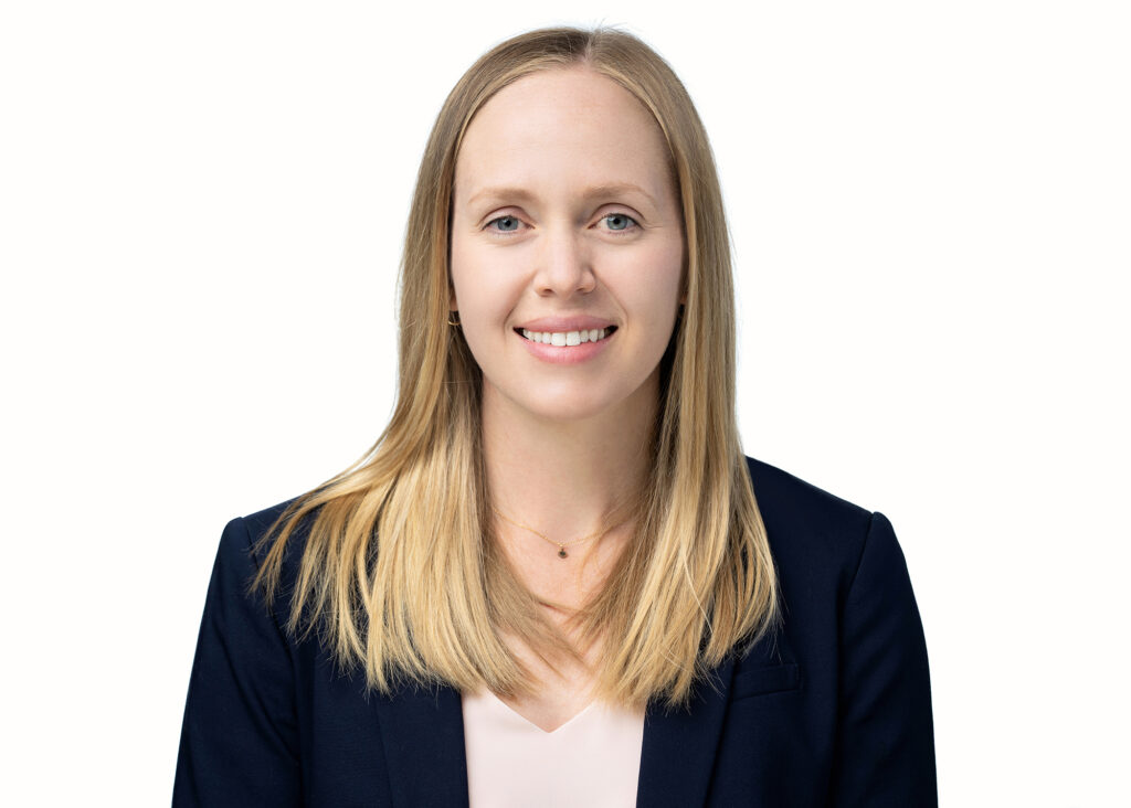 A woman smiles at the camera. She has blonde hair and is wearing a black jacket and standing in front of a plain white background. 