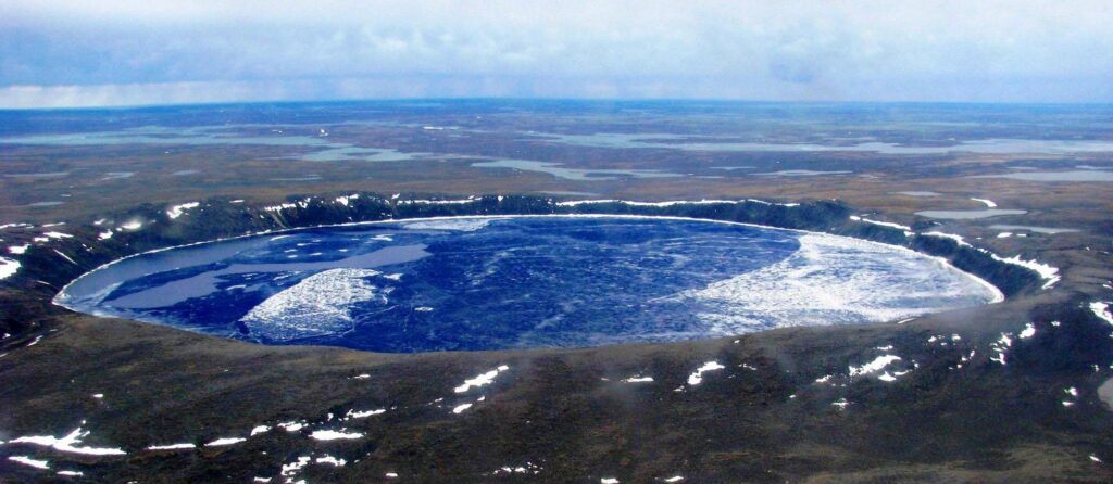 An aerial shot of the crater showing ice and water inside. The ground surrounding the crater is dark brown.
