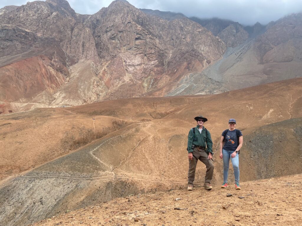 Two people standing in the middle ground looking at the camera. They are standing on a large red-ish plain and there is a red mountain range in the background.