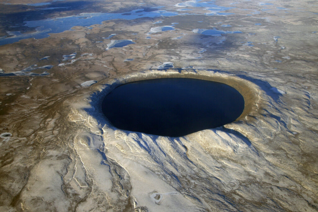 An aerial shot of the crater filled with dark water. The ground surrounding the crater is sandy-coloured.