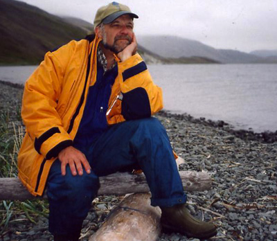 A man sits on a log with one knee up resting on a log in front of him. He is wearing a yellow jacket and hat and is resting his head in one hand. Behind him there is a lake and mountains