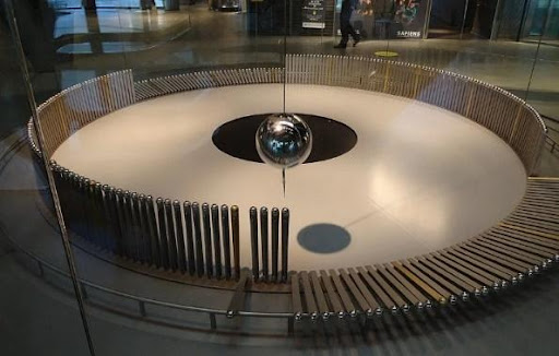 A large circle of standing small metal poles. In the middle of the circle a large ball is attached to a string and appears to be swinging back and forth, knocking over the poles. 