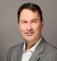 A man smiling at the camera. He is wearing a grey suit jacket and standing in front of a grey background