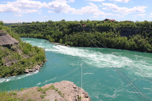 Aerial image of turquoise water with a whirlpool rapid in the middle of the river. There are cliffs on either side with trees and greenery.