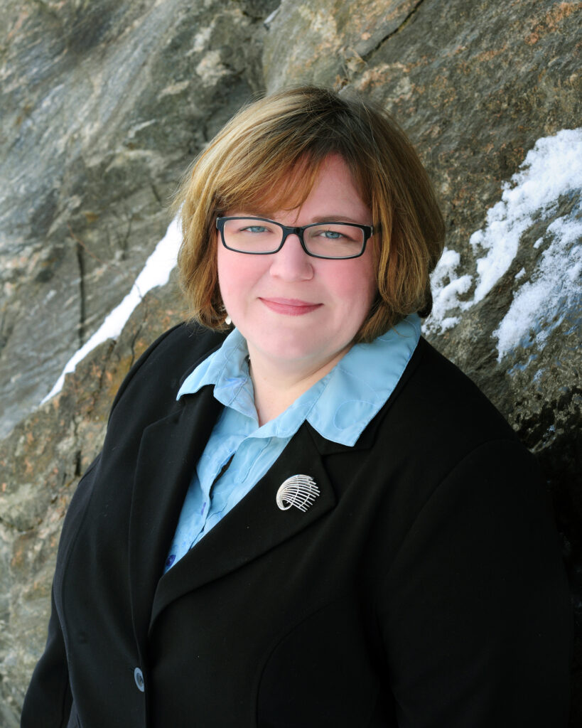 A woman smiles at the camera. She is standing in front of a steep cliff and is wearing a blue collared shirt and black sweater.