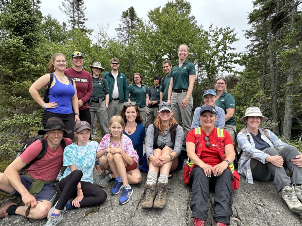 A group of 17 people stand or sit in two rows facing the camera. 7 of the people are dressed in green uniforms, and the others are dressed in a variety of colours. Joanna Hodge is in the front row wearing a high visibility jacket.