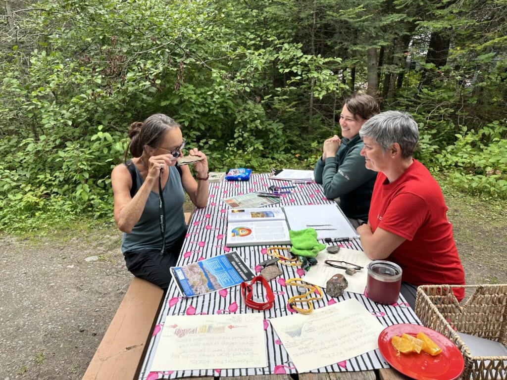 Three adults sit around a picnic table. On the table sit rocks, fossils, information sheets and hand lenses. One of the people is holding a rock to their eye and looking at it through a hand lens (magnifying glass).