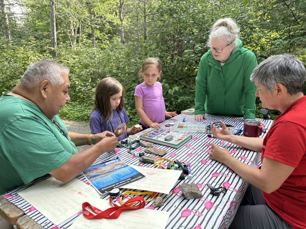 Three adults and two kids sit at a picnic table. On the table are rocks, fossils, information sheets, and hand lenses.