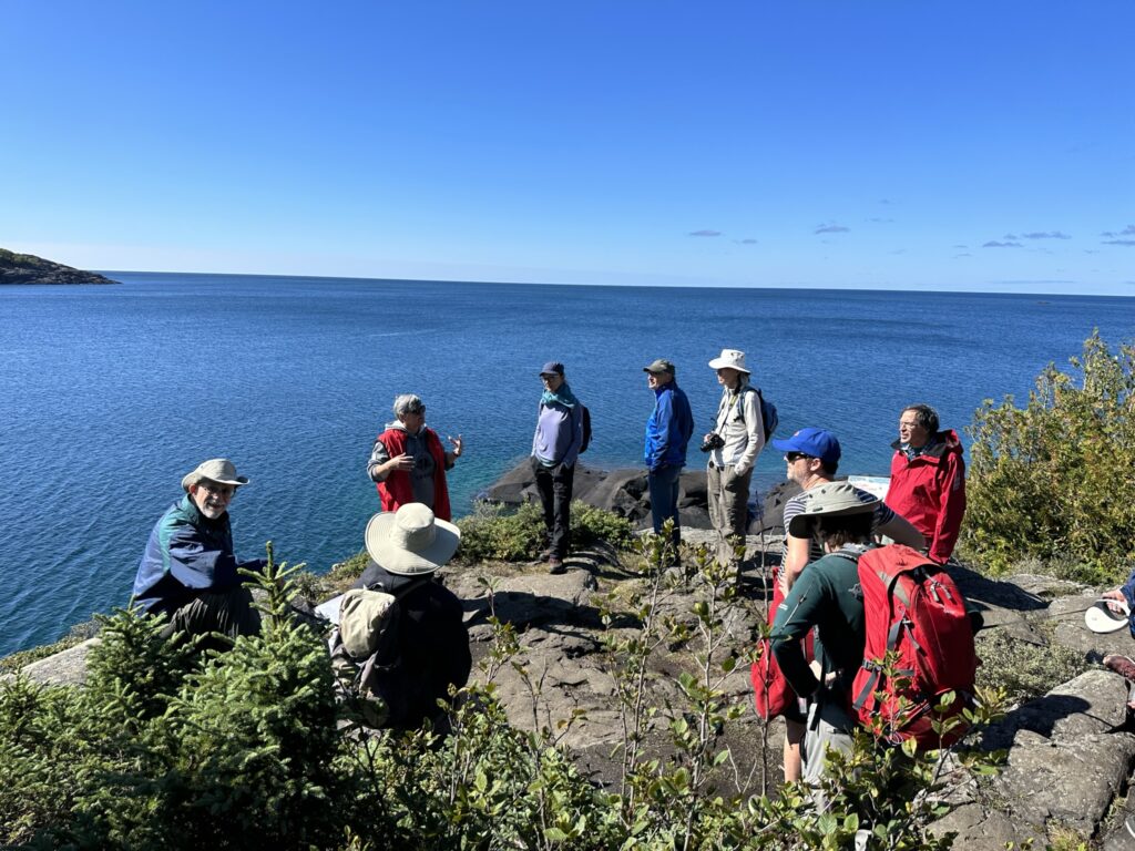 A group of 10 people stand or sit in a circle on a rock outcrop. Behind them water stretches to the horizon.