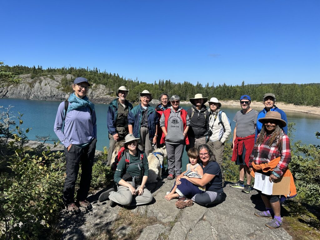Group of 13 people standing or sitting in a rough semicircle smiling at the camera. They are standing on a rock outcrop and behind them is a body of water with cliffs on the opposite side.