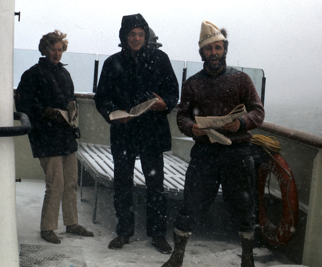 Three people standing on the deck of a boat with snow falling around them.