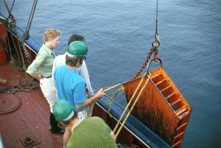 Four people stand on the deck of a boat watching a large orange device hanging off the side of the boat.